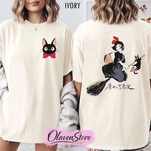 May include: Ivory-colored t-shirt featuring a black cat with a red bow tie on the front. The back of the shirt displays a cartoon illustration of a girl on a broom with a black cat, and Japanese text. The shirt has a crew neck.
