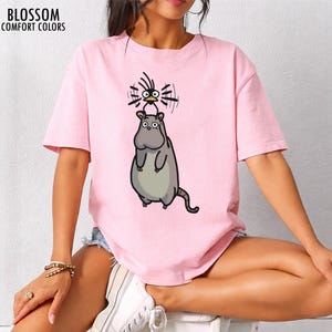 May include: A pale pink t-shirt with the text "BLOSSOM COMFORT COLORS" at the top left. The shirt features a cartoon illustration of a hippo-like creature with a bird on its head.