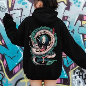 May include: Black hoodie featuring a colorful illustration of a dragon wrapped around a character with a mask. The dragon is white and teal, with a red box of Japanese text. The hoodie is worn by a person in front of a graffiti wall.