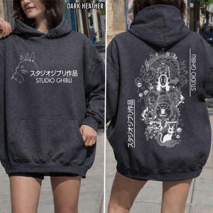 May include: Dark heather gray hoodie with a front kangaroo pocket. The front features a white graphic of a cartoon character and Japanese text. The back displays a detailed white illustration with Japanese text and the words "STUDIO GHIBLI".