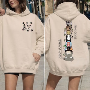 May include: Sand-colored hoodie with a front pocket and hood. The front features a pattern of black and white characters. The back displays a vertical stack of Studio Ghibli characters with Japanese text and the words "STUDIO GHIBLI".
