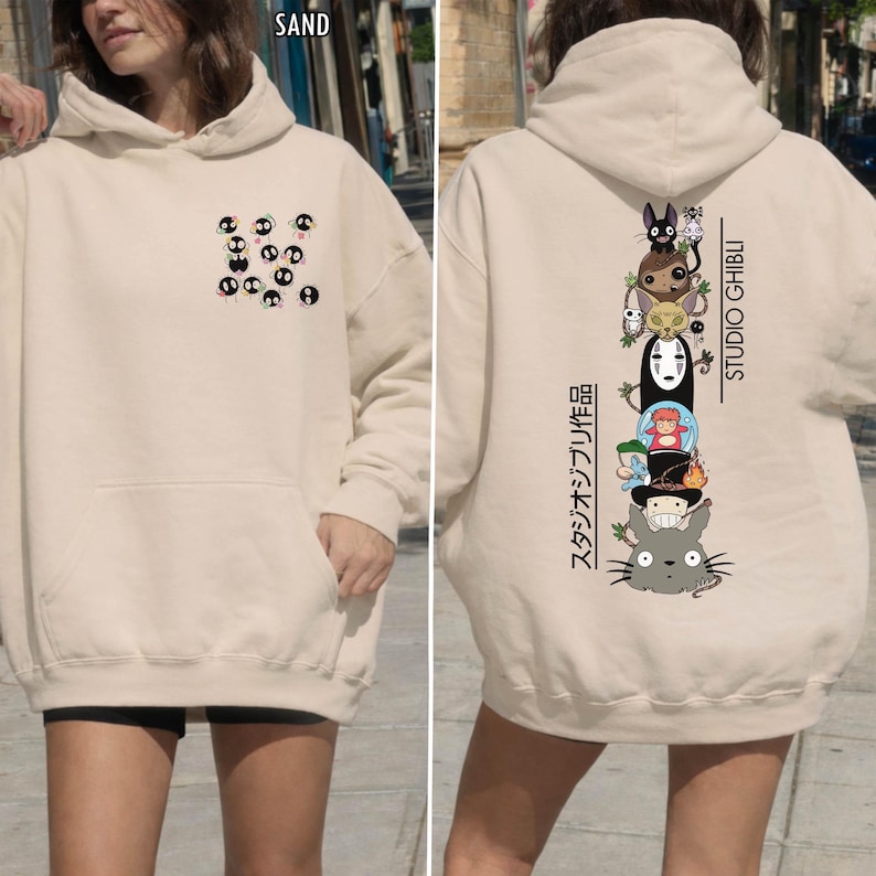 May include: Sand-colored hoodie with a front pocket and hood. The front features a pattern of black and white characters. The back displays a vertical stack of Studio Ghibli characters with Japanese text and the words "STUDIO GHIBLI".