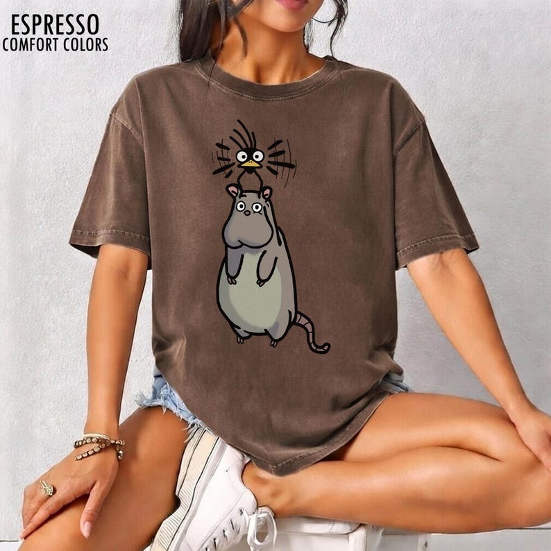 May include: Espresso-coloured Comfort Colors t-shirt with a cartoon hippo and bird design. The hippo is light grey with a long tail, and the bird is black with yellow eyes. The shirt has the words "ESPRESSO COMFORT COLORS" printed at the top.