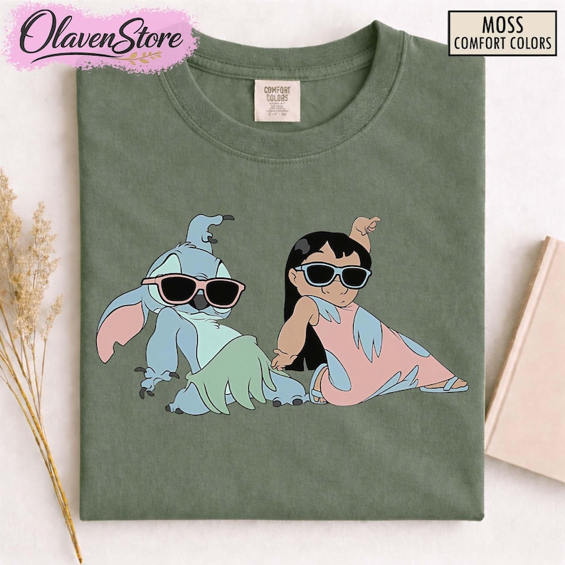 Op de afbeelding: Mosgroen Comfort Colors T-shirt met een afbeelding van Stitch en Lilo met een zonnebril. De stripfiguren zitten in een ontspannen pose, Stitch in blauw en Lilo in roze en blauw. Het shirt heeft de tekst "Moss Comfort Colors".