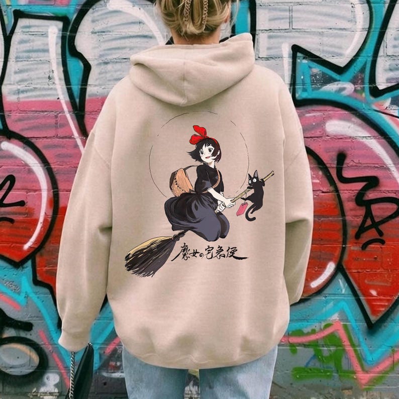 May include: Beige hoodie featuring a cartoon illustration of a young witch on a broom with a black cat. The witch wears a black dress and a red bow. The hoodie has a drawstring hood and long sleeves. The background is a colorful graffiti wall.