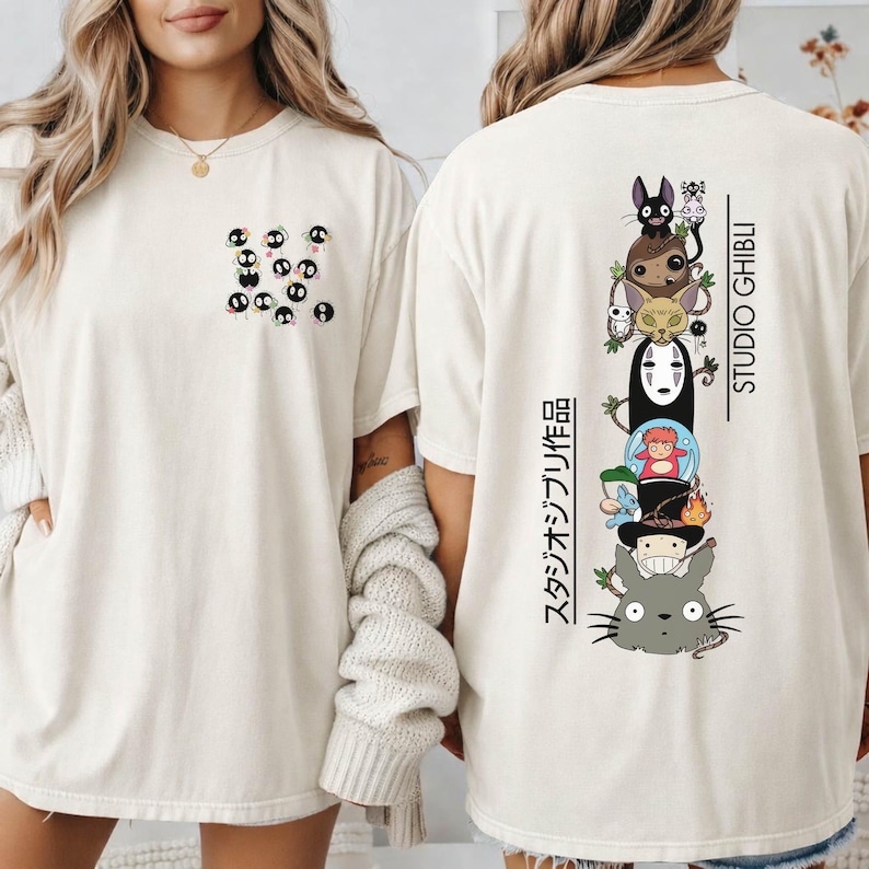 May include: Cream-colored t-shirt featuring a pocket design with black soot sprites. The back of the shirt displays a vertical stack of Studio Ghibli characters with Japanese text and the words "STUDIO GHIBLI" in black.