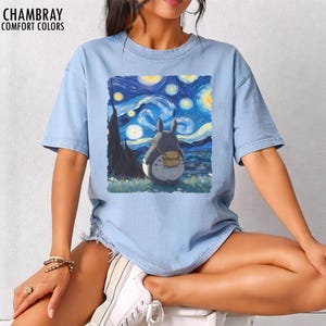 May include: Light blue t-shirt with a graphic of a cartoon character looking at a starry night sky, inspired by Van Gogh's "Starry Night." The shirt has the text "CHAMBRAY COMFORT COLORS" at the top left.
