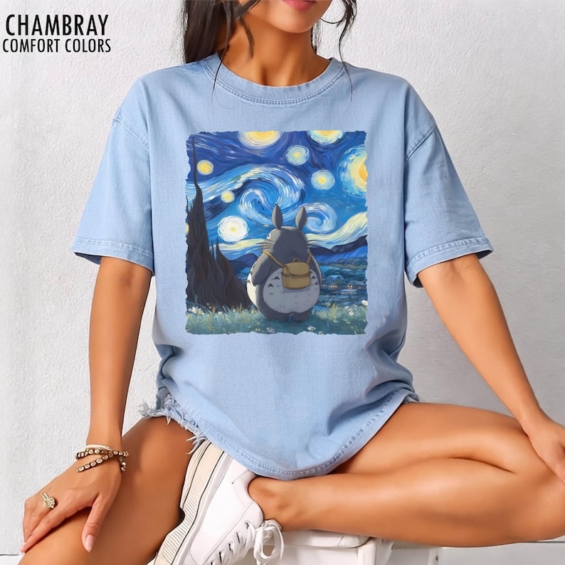 May include: Light blue t-shirt with a graphic of a cartoon character looking at a starry night sky, inspired by Van Gogh's "Starry Night." The shirt has the text "CHAMBRAY COMFORT COLORS" at the top left.