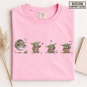 May include: Pink t-shirt with a graphic of a green alien-like character in various poses. The character is shown in a pod, playing with a ball, confused, and saying "coo!". The shirt has the text "BLOSSOM COMFORT COLORS".