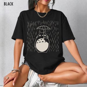 May include: Black t-shirt featuring a white illustration of a cartoon character holding an umbrella in the rain. The text "BLACK" is at the top left of the shirt. The design is a simple line drawing style.