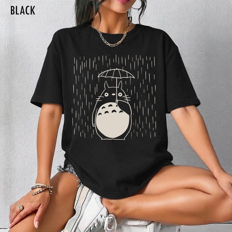 May include: Black t-shirt featuring a white illustration of a cartoon character holding an umbrella in the rain. The text "BLACK" is at the top left of the shirt. The design is a simple line drawing style.