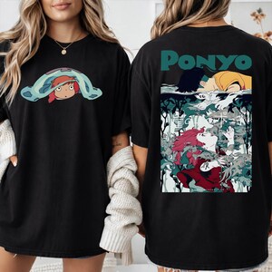 May include: Black t-shirt featuring a cartoon graphic of a character under a blue blanket on the front. The back of the shirt has a graphic with the word "PONYO" and an illustration of a girl with red hair.