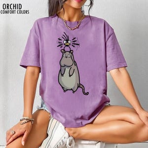 May include: A lilac-coloured t-shirt from Orchid Comfort Colors, with a cartoon hippo and bird design. The hippo is grey, and the bird is black and yellow. The text "ORCHID COMFORT COLORS" is at the top left of the shirt.