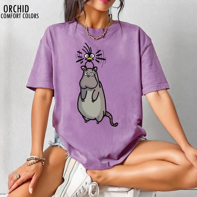 May include: A lilac-coloured t-shirt from Orchid Comfort Colors, with a cartoon hippo and bird design. The hippo is grey, and the bird is black and yellow. The text "ORCHID COMFORT COLORS" is at the top left of the shirt.