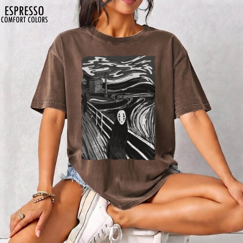 Op de afbeelding: Espresso-kleurig t-shirt met een zwart-witte grafische print. De afbeelding toont een gestileerde sc&egrave;ne met een figuur in een lange zwarte mantel en een wit masker, die doet denken aan een bekend schilderij. De tekst "ESPRESSO COMFORT COLORS" staat bovenaan.