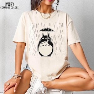 May include: Ivory-colored t-shirt featuring a black graphic of a cartoon character holding an umbrella in the rain. The text "IVORY COMFORT COLORS" is printed at the top left. The shirt is a short-sleeved, casual style.