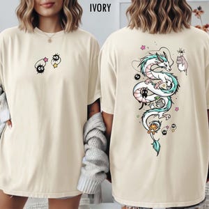 Pode incluir: Camiseta de cor marfim com um design extravagante. A frente apresenta duas personagens de desenhos animados, enquanto a parte de tr&aacute;s exibe uma ilustra&ccedil;&atilde;o detalhada de um drag&atilde;o com detalhes azuis e brancos, acompanhada por outras figuras animadas. Uma pe&ccedil;a de roupa confort&aacute;vel e casual.