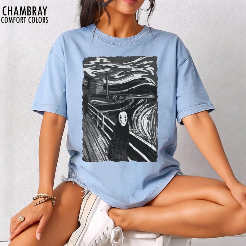 Op de afbeelding: Lichtblauw T-shirt met een zwart-witte grafische print. De afbeelding toont een gestileerde versie van het schilderij "De Schreeuw" met een personage uit de film "Spirited Away". De tekst "CHAMBRAY COMFORT COLORS" staat bovenaan.