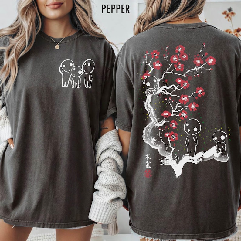 May include: Pepper-colored t-shirt featuring a white graphic of three figures on the front. The back displays a tree with red flowers, additional figures, and Japanese text. The shirt has a relaxed fit.