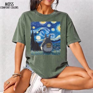 May include: Olive green t-shirt with a graphic of Totoro from My Neighbor Totoro, set against a backdrop inspired by Van Gogh's "Starry Night." The shirt features the text "MOSS COMFORT COLORS" at the top left.