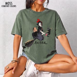 May include: Moss Comfort Colors olive green t-shirt featuring a cartoon illustration of a witch riding a broom with a black cat. The witch wears a red bow and black dress. Japanese text is below the illustration.