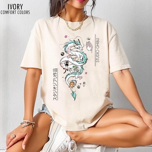 May include: Ivory-colored t-shirt with a graphic of a white dragon with teal accents, surrounded by cartoon characters. The text "STUDIO GHIBLI" and Japanese text are printed vertically on the shirt. The shirt is labeled "IVORY COMFORT COLORS" in the upper left corner.