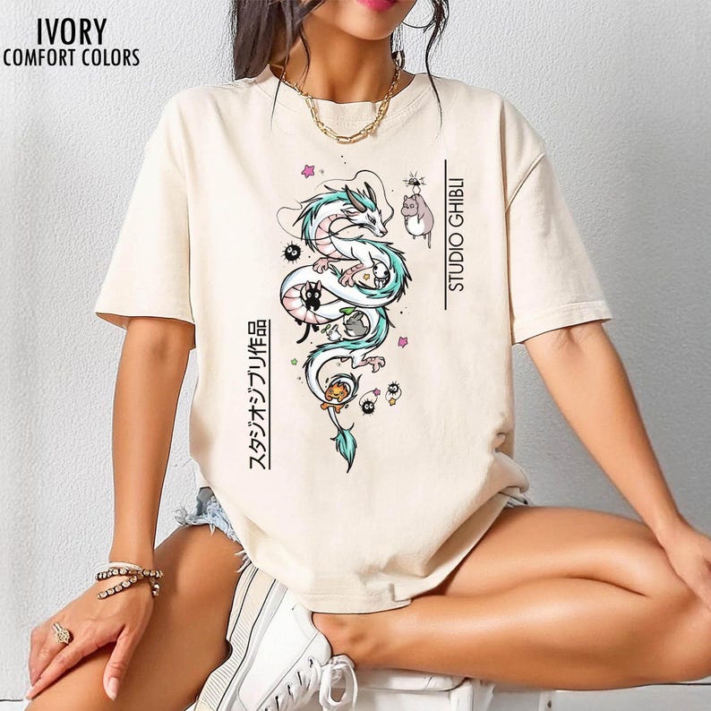 May include: Ivory-colored t-shirt with a graphic of a white dragon with teal accents, surrounded by cartoon characters. The text "STUDIO GHIBLI" and Japanese text are printed vertically on the shirt. The shirt is labeled "IVORY COMFORT COLORS" in the upper left corner.