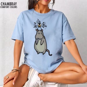 May include: Pale blue t-shirt featuring a cartoon hippo and bird design. The hippo is grey with a green belly and a long tail. The bird is black with a spiky head. The text "CHAMBRAY COMFORT COLORS" is at the top left of the shirt.