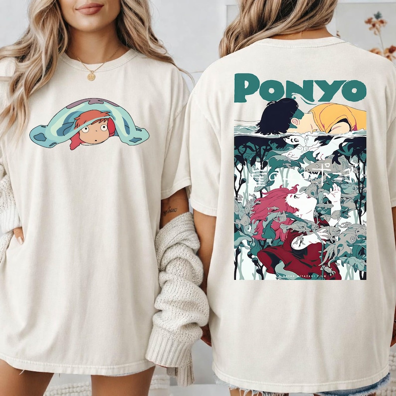 May include: Off-white t-shirts with a cartoon graphic of a character from the animated film Ponyo. The front of the shirt features a character peeking out from under a blanket. The back of the shirt has the word "Ponyo" and a scene from the film.