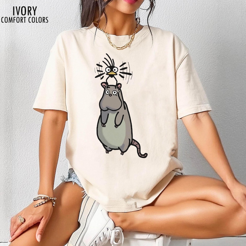 May include: Ivory t-shirt featuring a cartoon hippo-like character with a bird perched on its head. The text "IVORY COMFORT COLORS" is printed at the top left. The shirt has short sleeves and a loose fit, suitable for casual wear.
