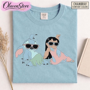 Op de afbeelding: Lichtblauw T-shirt met een afbeelding van Stitch en Lilo van Lilo & Stitch, beiden met een zonnebril. Het shirt heeft de tekst "CHAMBRAY COMFORT COLORS" in een zwarte doos. De merknaam "OlavenStore" staat linksboven.