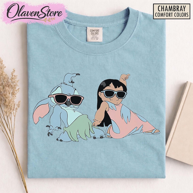 Op de afbeelding: Lichtblauw T-shirt met een afbeelding van Stitch en Lilo van Lilo & Stitch, beiden met een zonnebril. Het shirt heeft de tekst "CHAMBRAY COMFORT COLORS" in een zwarte doos. De merknaam "OlavenStore" staat linksboven.