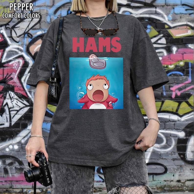 May include: Dark grey t-shirt featuring the word "HAMS" in red above a cartoon image of a character in water. The character is red and has a surprised expression. The shirt has the words "PEPPER COMFORT COLORS" in the upper left corner.