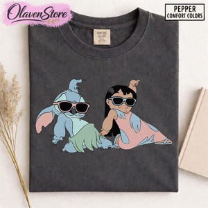Op de afbeelding: Donkergrijs t-shirt met een afbeelding van twee stripfiguren met een zonnebril. E&eacute;n is blauw met roze oren en de ander is een meisje in een roze jurk. Het shirt heeft de tekst "Pepper Comfort Colors".
