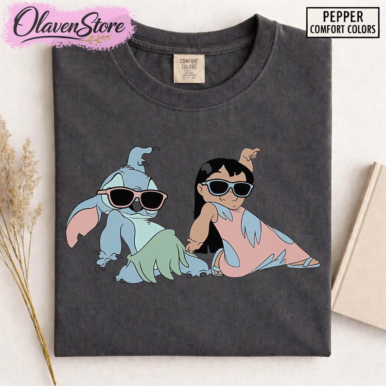 Op de afbeelding: Donkergrijs t-shirt met een afbeelding van twee stripfiguren met een zonnebril. E&eacute;n is blauw met roze oren en de ander is een meisje in een roze jurk. Het shirt heeft de tekst "Pepper Comfort Colors".