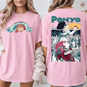 May include: Pink t-shirt featuring a cartoon graphic of a character from the animated film "Ponyo" on the front and back. The front graphic shows the character under a blue blanket. The back graphic has the word "Ponyo" and a scene from the film.