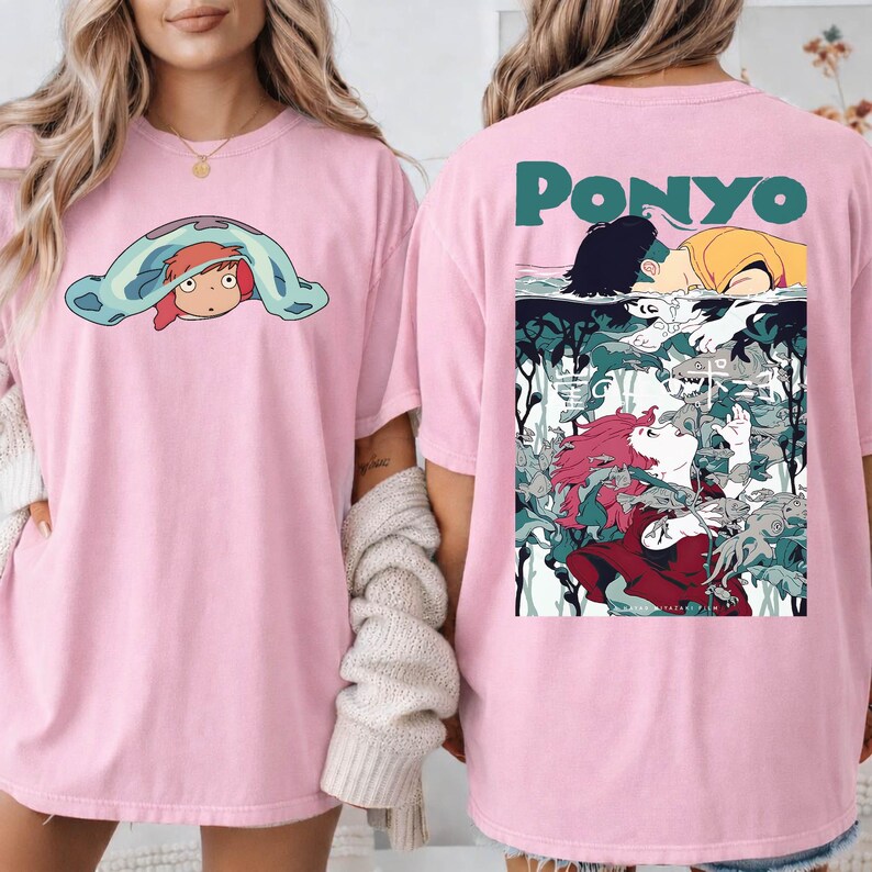 May include: Pink t-shirt featuring a cartoon graphic of a character from the animated film "Ponyo" on the front and back. The front graphic shows the character under a blue blanket. The back graphic has the word "Ponyo" and a scene from the film.