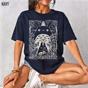 以下が含まれることがあります： 紺色のTシャツ。クリーム色のグラフィックで、アニメ映画「となりのトトロ」のキャラクターが森のシーンに描かれています。「NAVY」の文字が左上にプリントされています。