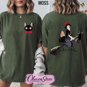 May include: Moss green t-shirt featuring a black cat with a red bow tie on the front. The back of the shirt displays a graphic of a girl on a broom with a black cat. The text "魔女の宅急便" is also on the back. The shirt has a relaxed fit.