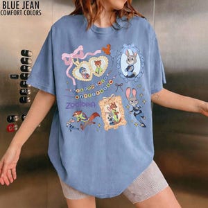 May include: A blue jean colored t-shirt featuring a graphic print of Zootopia characters including Judy Hopps and Nick Wilde. The design includes ornate frames, a pink bow, and the text Welcome to Zootopia and Zootopia in purple lettering.