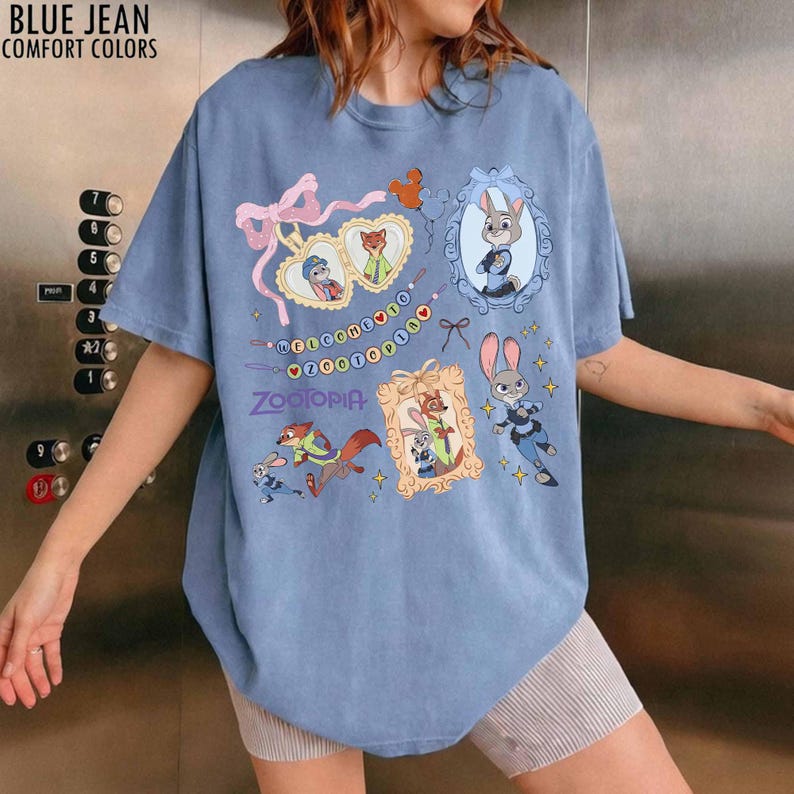 May include: A blue jean colored t-shirt featuring a graphic print of Zootopia characters including Judy Hopps and Nick Wilde. The design includes ornate frames, a pink bow, and the text Welcome to Zootopia and Zootopia in purple lettering.