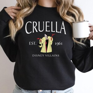 May include: Black sweatshirt with the word "CRUELLA" in white arched text above an illustration of Cruella de Vil. The text "EST. 1961" and "DISNEY VILLAINS" are also printed on the sweatshirt.