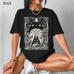 以下が含まれることがあります： 黒いTシャツ。クリーム色のグラフィックで、映画「となりのトトロ」のキャラクターが森の中に描かれています。デザインにはトトロ、蝶、葉が含まれています。「BLACK」の文字が上部に印刷されています。