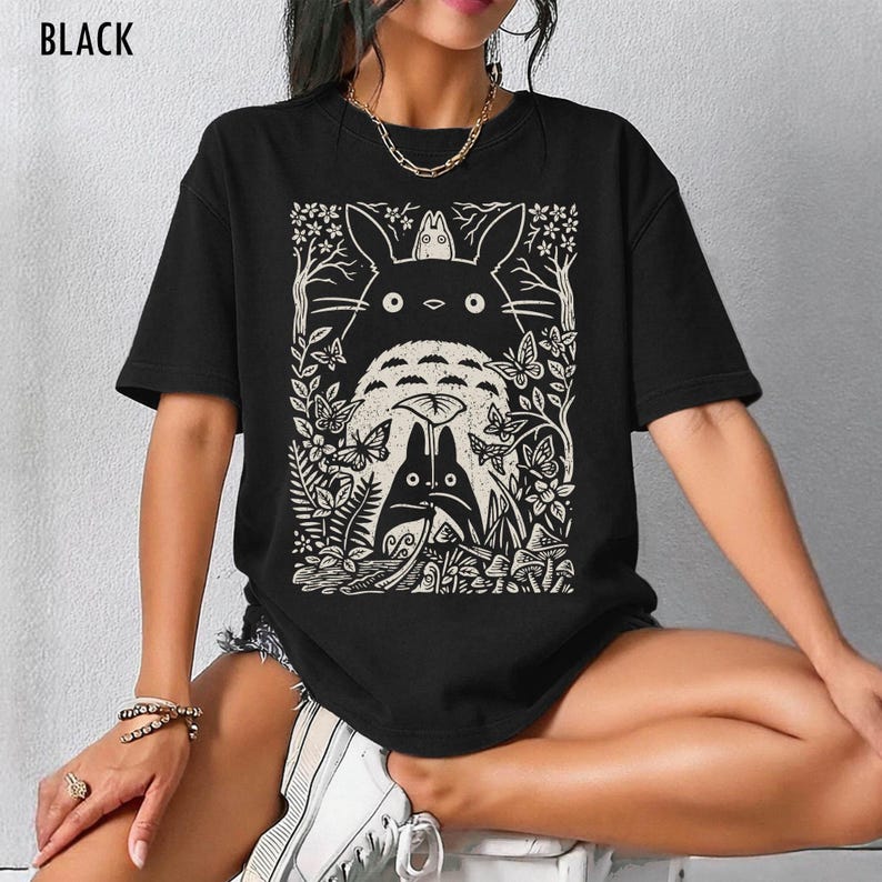 以下が含まれることがあります： 黒いTシャツ。クリーム色のグラフィックで、映画「となりのトトロ」のキャラクターが森の中に描かれています。デザインにはトトロ、蝶、葉が含まれています。「BLACK」の文字が上部に印刷されています。