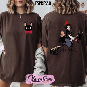 May include: Espresso-colored t-shirt featuring a black cat with a red bow tie on the front. The back of the shirt displays a graphic of a witch on a broom with a black cat and Japanese text. The shirt is from OlavenStore.