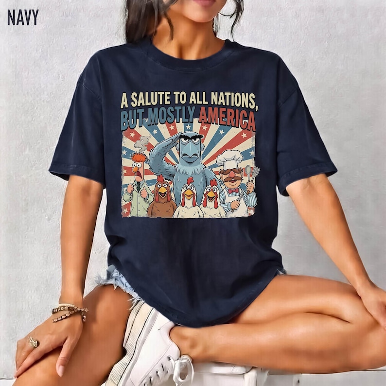 May include: Navy blue t-shirt featuring a graphic with the text "A SALUTE TO ALL NATIONS, BUT MOSTLY AMERICA." The graphic includes cartoon characters, an eagle, and a patriotic background.