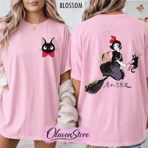 May include: Light pink t-shirt featuring a black cat with a red bow tie on the front and a graphic of a witch on a broom with a black cat on the back. The word "BLOSSOM" is printed at the top.