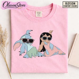 Op de afbeelding: Roze T-shirt met een cartoon illustratie van Stitch en Lilo met een zonnebril. De personages zitten in een ontspannen pose. Het shirt heeft de tekst "Blossom Comfort Colors".