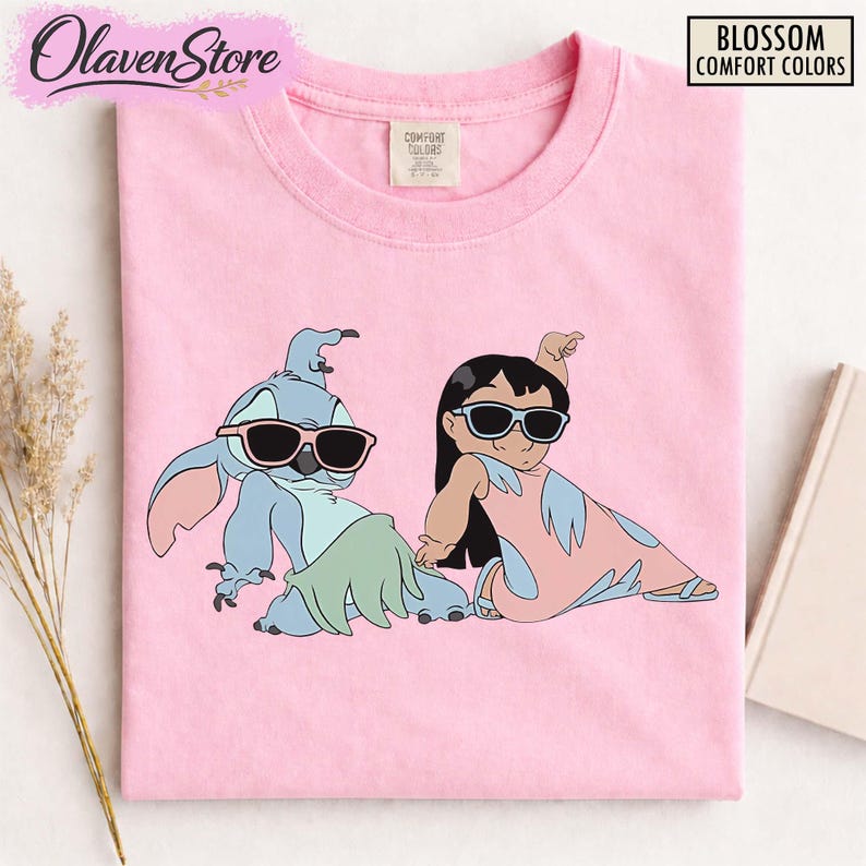 Op de afbeelding: Roze T-shirt met een cartoon illustratie van Stitch en Lilo met een zonnebril. De personages zitten in een ontspannen pose. Het shirt heeft de tekst "Blossom Comfort Colors".