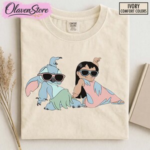 Op de afbeelding: Ivoor Comfort Colors T-shirt met een cartoon illustratie van Stitch en Lilo met een zonnebril. Het shirt heeft het OlavenStore logo en de woorden "IVORY COMFORT COLORS" in een kader. Een casual, leuk ontwerp.